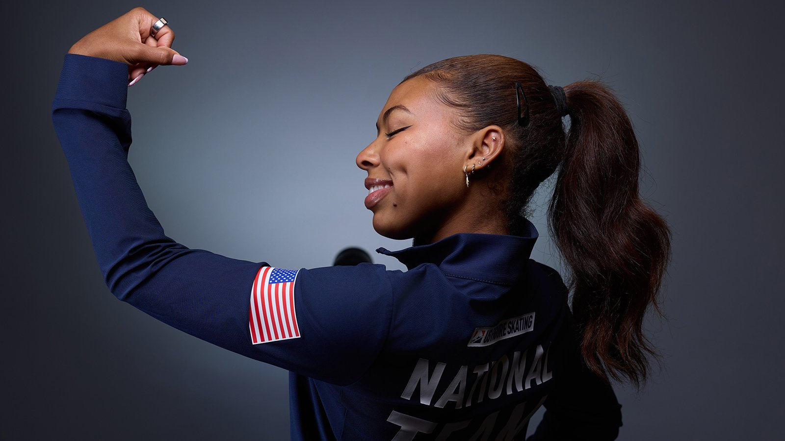 With back turned, Starr Andrews shows her muscle with an arm flex and a smile. She's wearing a U.S. National Team jacket.