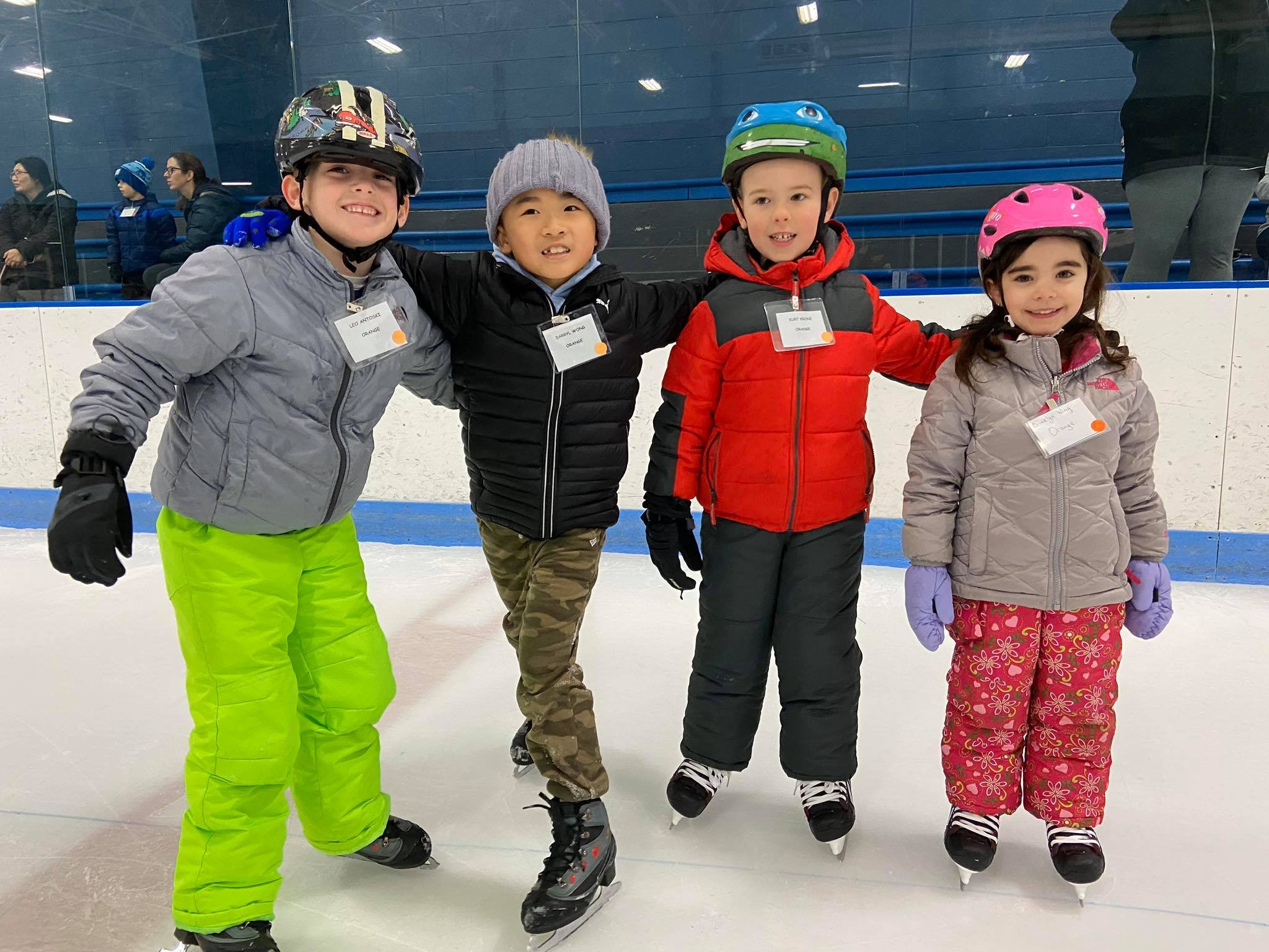 Four young skaters pose on the ice. They are all wearing multi-colored winter jackets and ski pants