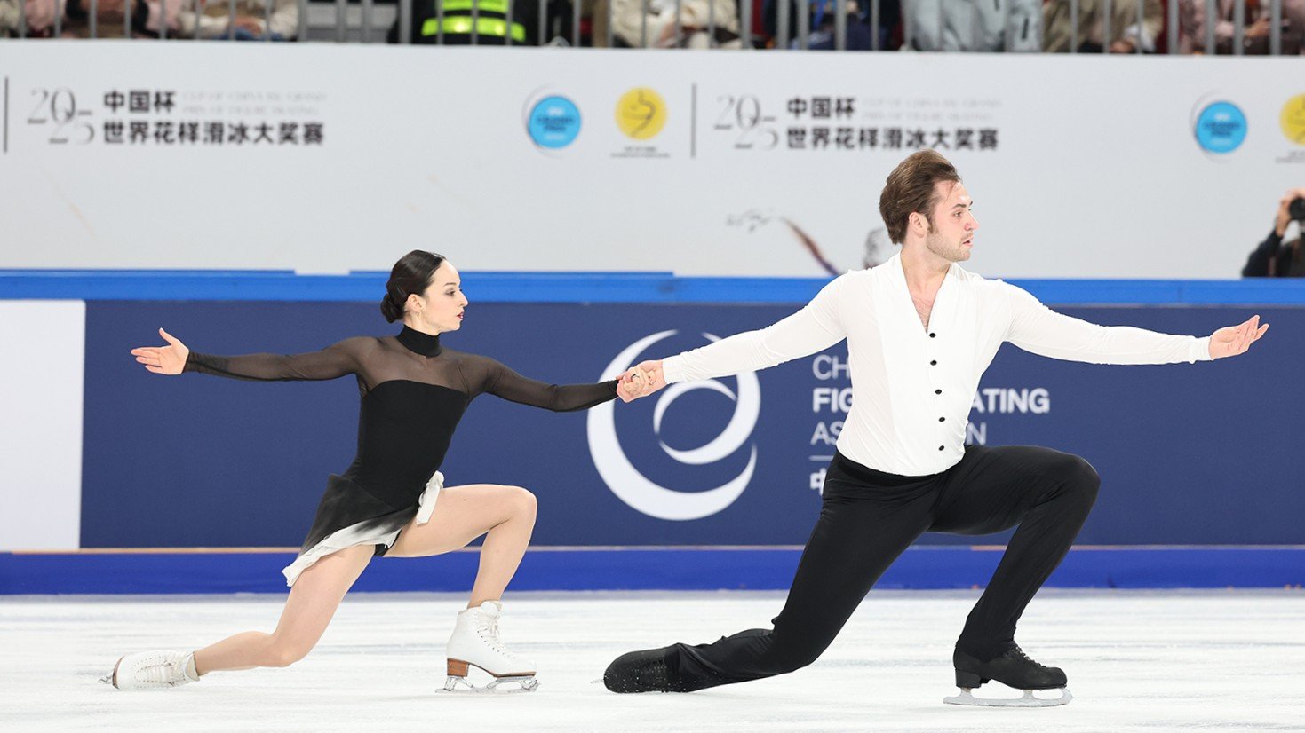 Katie McBeath and Daniil Parkman kneel in unison while holding hands. Katie (left) is a young woman with black hair tied back in a low bum wearing a black skating costume with sheer long sleeves. Daniil (right) is a man with short brown hair wearing a long sleeve white button down top and black pants