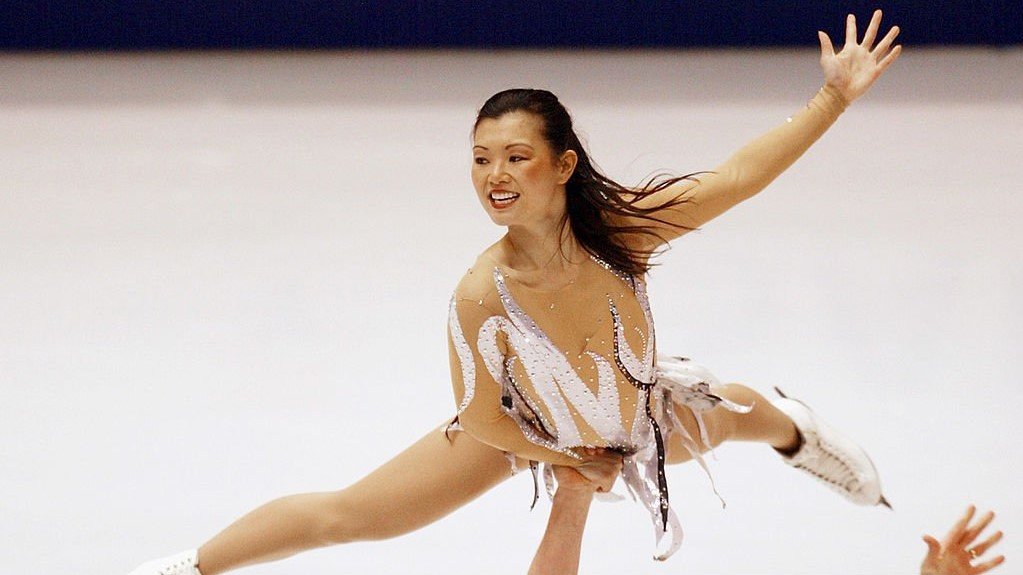 Kyoko Ina is held up above her partners head in a lift while performing at the 2002 Winter Olympic Games in Salt Lake City. Kyoko is a young Asian woman with long black hair. She is wearing a white skating costume and white skates. 