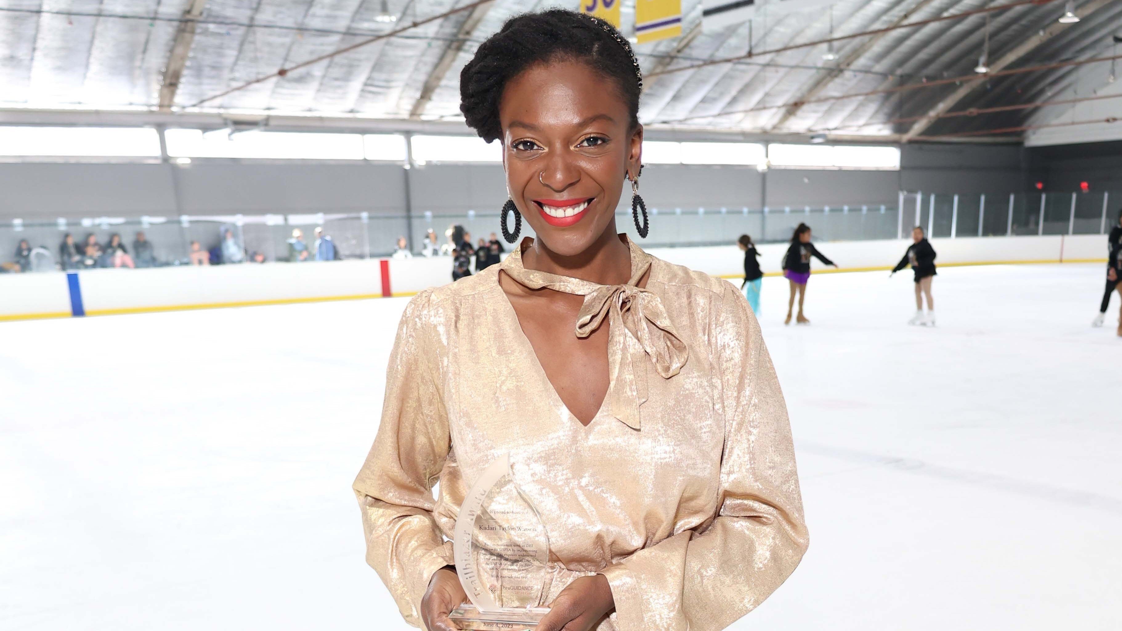 Kadari Taylor-Watson poses on the ice with her Trailblazer Award. She is a Black woman with her hair up. She is wearing large black curcular earrings and a gold top with a bow around the neck. She is holding a glass award which is in the shape of a tear drop