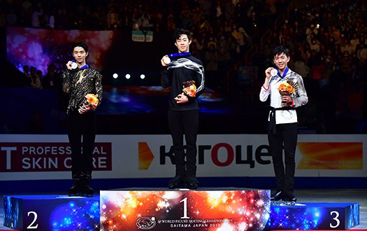 Nathan Chen stands atop the podium, with Yuzuru Hanyu to his right, and teammate Vincent Zhou to his left, hold his medal towards the camera.