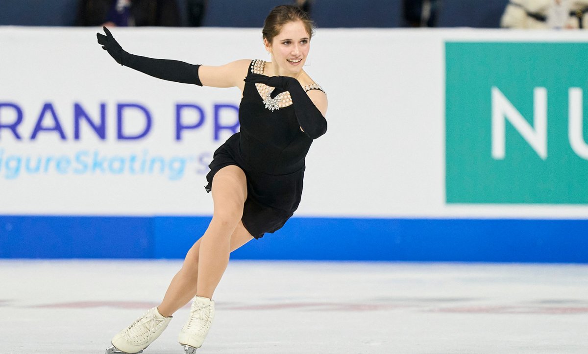 Isabeau Levito, wearing a black dress with pearl embellishment at the top and black gloves, skates to the right, a smile on her face.