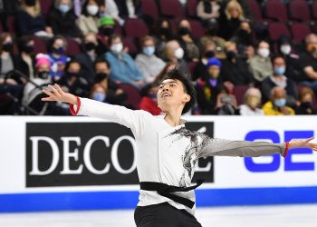 Vincent Zhou looks to the sky with his arms outstretched while on the ice and wearing a black and white costume.