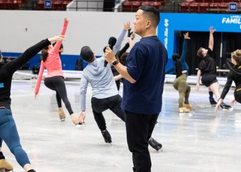 Gig stands in the forefront of the photo with a mic in his hand, facing skaters who are practicing Disney on Ice  choreography.