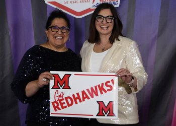 Standing up against the 2024 U.S. Synchronized Skating Championships kiss and cry backdrop, Carla DeGirolamo and LeeAnn Shoker hold up a Redhawks sign. Both wear sequins.