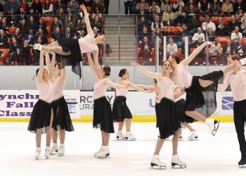 The Haydenettes perform a lift with two skaters hoisted in the air. They are all wearing matching skating costumes with a light pink top and black skirt. One male skater is wearing a coordinating top with black pants.