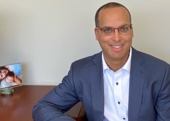 Aaron Parchem is an African American man wearing a blue suit with a white collared shirt underneath and glasses. He sits at a desk with his right elbow on the table. A photo of his young kids sits on the desk behind him