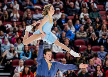 Alisa Efimova and Misha Mitrofanov complete a lift with Alisa being lifted over Misha's head. Alisa is wearing a light blue skating costume and Misha is wearing a coordinating dark blue long sleeve top
