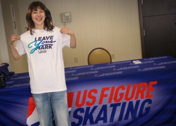 Alysa Liu smiles wearing a white tshirt with the Leave Your Mark logo. She stands in front of a table with a U.S. Figure Skating branded tablecloth on top.