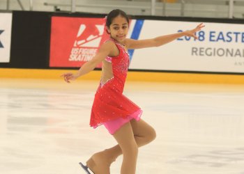 Photo of Anya performing on the ice. She is a young South Asian girl with black hair in a low bun. She is wearing a pink skating costume and she is standing with her arms outstretched. 