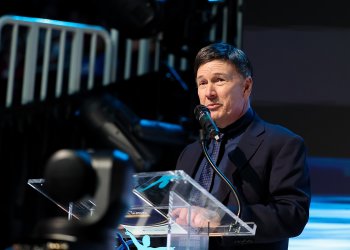 U.S. Figure Skating interim CEO Sam Auxier stands at a clear podium speaking to the crowd. Sam is an older man with short brown air wearing a blank button down shirt, black suit jacket and a navy blue tie. 