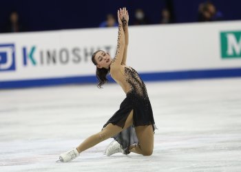 Ava Ziegler kneels on the ice with both arms raised above her head. She is a young woman with long brown hair wearing a black and tan skating costume. 