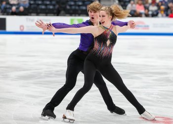 Emily Bratti and Ian Somerville during their 80s themed rhythm dance. She wears a blacktop with a colorful detail and black pants while he wears black pants and a purple top. As they skate backwards, both have an excited, energetic expression.