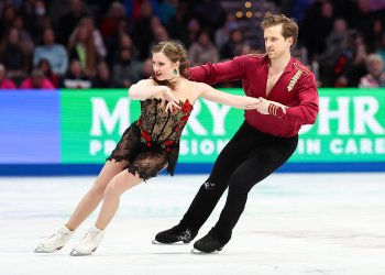 Christina Carreira and Anthony Ponomarenko skate together with Anthony skating behind Christina, holding on to her outstretched arm. Anthony is a young man wearing a long sleeve red top and Christina is a young woman with long brown hair wearing a black lace skating costume with red rose details. 
