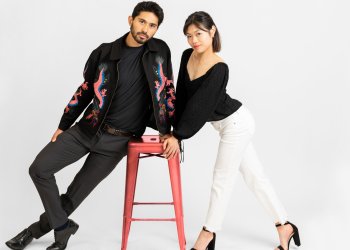 Emily Chan and Spencer Howe learn on a stool while posing for their cover shoot for SKATING magazine. Howe (left) is a man with short black hair wearing a black jacket with a bright pink and blue dragon design over a black shirt and black jeans. Chan is a woman with short black hair wearing a black long sleeve shirt, white jeans, and black high heels 