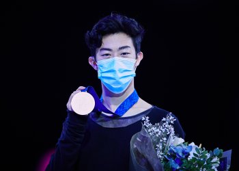 Nathan Chen holds up his 2021 Worlds gold medal while wearing a mask and holding flowers in dark lighting.