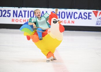Katarina Suemnig rides an inflatable yellow chicken. The 2025 National Showcase banner is in the background. She's wearing a green costume with padding. 