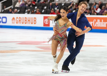 Madison Chock and Evan Bates compete in the rhythm dance at the 2023 Toyota U.S. Figure Skating Championships. Madison (left) is a woman with long black hair wearing a green, pink and purple sparkly skating costume. She has a big smile and her arms are extended outward. Evan skates behind her holding her hand and waist. He is a white man with short blonde hair wearing a blue top with a deep v neck and matching blue pants. 