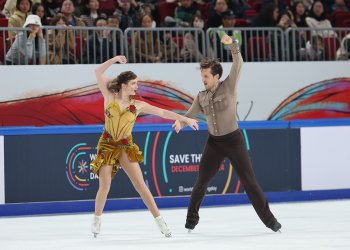 Facing each other, hand in hand, Christina Carreira and Anthony Ponomarenko perform the "Carmen" free dance. Christina is a young woman with long brown hair wearing a yellow skating costume with red flowers. Anthony is a young man with shirt brown hair wearing a light brown button down long sleeve top and brown pants