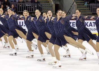 DC edge skates together in parallel lines with their arms around each other. They are all wearing matching navy blue skating costumes with sheer sleeves and silver stripes on the top. 