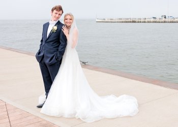 Logan Bye (left) in a navy suit and Eva Pate (right), in a wedding dress and veil. They stand in front of a lake and marina of boats.