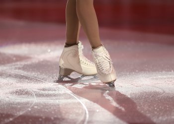 Photo of a skaters legs on the ice wearing white skates. A red light shines over the ice. 