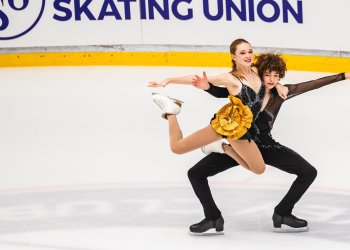 Olivia Ilin and Dylan Cain compete at JGP Hungary. Ilin is a young woman with long blonde hair and Cain is a young man with curly brown hair. Cain holds up Ilin in a lift. Cain is wearing a long sleeve black top with black pants and Ilin is wearing a black skating costume with a gold bow detail at the hip