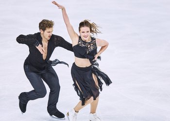 Christina Carreira and Anthony Ponomarenko on the ice. Both wear black. He skates behind her, her holding her waist. She holds his hand on her waist, her other arm up in the air.