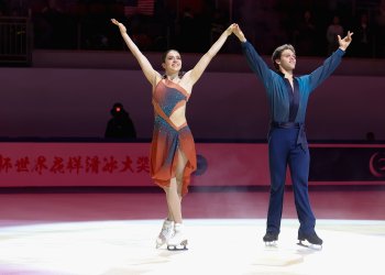 Caroline Green and Michael Parsons salute the crowd after their free skate. They stand in the spotlight, smiling with their arms in the air as they look into the crowd. 