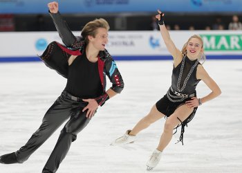  Artem Markelov and Leah Neset skate toward the camera during their rhythm dance. They have the same movement, their right arm up in the air and left arm on their hip. She wears a leather dress with big bold silver jewelry. He wears black leather pants and a black shirt with a black lather jacket that has red and light blue detail.