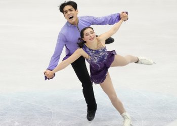Olivia Flores and Luke Wang compete in the short program at the ISU World Figure Skating Championships 2024. Olivia skates in front of Luke wearing a dark purple skating costume. She has one leg extended behind her and her arms are outstretched to hold Luke's hands. Luke skates behind her wearing a long sleeve light purple top and black pants. He is holding both of Olivia's hands.