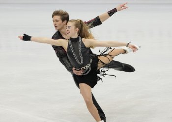 Leah Neset and Artem Markelov skate together with one leg extended behind them. Neset has both arms outstretched and Markelov skates beside Neset with one hand on her waist and the other extended outward. They are both wearing black leather costumes.