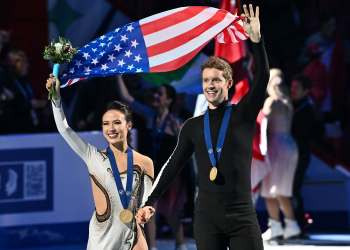 Madison chck and Evan bates hold hands, gold medals around their necks. With their outside arm, they wave to the crowd, holding the U.S. flag in the air behind them.