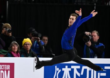 Dressed in a blue top and black pants, Jason Brown soars through the air with legs spread and arms extended.