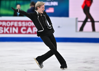 Ilia Malinin, in a black shirt, jacket, pants and gloves, looks to the right, his left hand out, a fierce expression on his face.