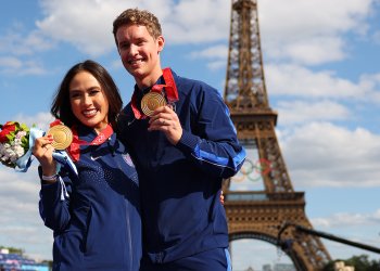 Madison Chock and Evan Bates, wearing blue Nike jackets, hold up their gold medals in Paris, when they received their Olympic gold medals for the 2022 team event.