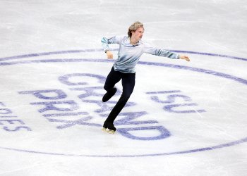 Ilia Malinin, dressed in a white top and black pants, skates over the ISU Grand Prix logo at Skate Canada International.