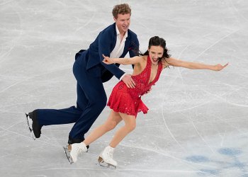 Madison Chock and Evan Bates compete in the rhythm dance at 2025 World Team Trophy. Madison is a woman with long black hair wearing a red skating costume with a deep v neck. She skates with her arms out and an excited look on her face. Evan skates behind her holding her waist. Evan is a tall man with shirt curly blonde hair wearing a navy blue suit with a white button down shirt underneath. 