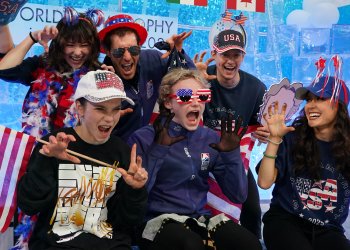 Team USA makes a funny face at the camera while sitting in the team box at World Team Trophy. Ilia sits in the middle wearing his team jacket while everyone else wears various patriotic gear and waves USA flags. 