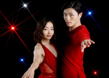 Maia Shibutani (left) and Alex Shibutani (right) stand in front of a black background with red, white and blue lights facing each other with their arms extended. Maia is a young woman with short shoulder length black hair wearing a sleeveless red skating costume. Alex is a young man with short black hair wearing a matching red long sleeve top. 