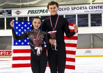 RIGA, LATVIA - AUGUST 22: Bronze medal winners Naomi Williams and Lachlan Lewer of the United States pose with their medals during medal ceremony after the ISU Junior Grand Prix of Figure Skating at Volvo Sporta Centrs on August 22, 2025 in Riga, Latvia. They stand on the ice, holding the U.S. flag behind them and holding a bouquet of flowers. Both wear black skating costumes.