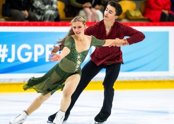 Jasmine Robertson and Chase Rohner skate together. Jasmine skates in front. She is a young woman with long blonde hair wearing a green skating costume with a v neck. Chase skates behind Jasmine holding on to her left arm. Chase is a young man with short brown hair wearing a long sleeve dark red top with a deep v neck and black pants 