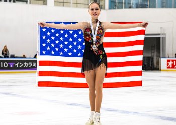 Sophie Joline von Felten stands proudly, smiling while holding a United States flag behind her with a bronze medal with a white ribbon around her neck. Sophie Joline is a young woman with blonde hair tied back in a bun wearing a black skating costume with multicolored flower detailing down the front and sheer long sleeves. 