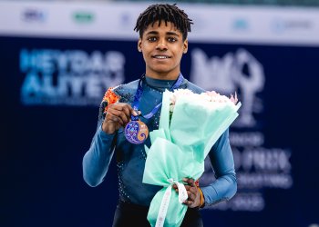 Caleb Farrington smiles while holding his bronze medal in one hand and a bouquet of flowers in the other. Caleb is a young Black man with braces and short dreadlocks wearing a dark teal top with orange detailing on the shoulder and black pants. 