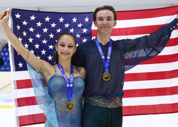 Hana Maria Aboian and Daniil Veselukhin stand smiling with gold medals around their necks. They are both holding the corners of an American flag behind them. Hana (left) is a young woman with long brown hair tied back in a ponytail wearing a blue skating costume with spaghetti straps and blue fabric resembling wings down the side. Daniil (right) is a tall young man with short brown hair wearing a dark blue long sleeve top with sheer sleeves and a beaded belt. 