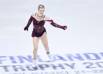 Amber Glenn, in a maroon dress with cold shoulder sleeves and her hair up, skates at center ice at 2025 Finlandia Trophy. She smiles as she skates.