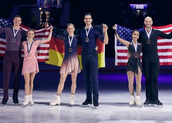 The 3 pairs teams who medaled at Finlandia Trophy. To the left, Alisa Efimova and Misha Mitrofanov, in the middle the German team and to the right Ellie Kam and Danny O'Shea. The two U.S. pairs teams stand holding U.S. flags behind them, the Germans hold their flag behind them. All smile with medals around their necks.