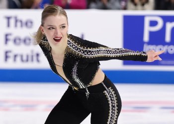 Amber Glenn skates toward the camera, her hair pulled back in a pony tail. The picture is close up, taken from her thigh up. She wears a black bodysuit with silver and gold jewels.