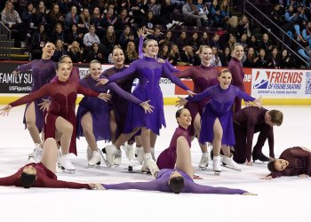 The Haydenettes strike their ending pose with smiles on their face. The team is wearing coordinating purple or red long sleeve skating costumes.